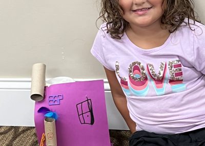 A young girl is seated on the floor, engaged with a handmade paper house, showcasing her creativity and imagination.