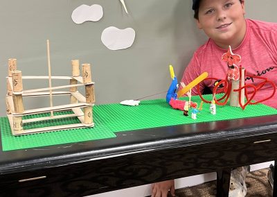 A boy sits on the floor next to a table that shows what he has created with crafting materials like corks, popsicle sticks, and pipe cleaners.