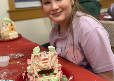 A girl with a reindeer antler headband smiles in front of a decorated gingerbread house on a red tablecloth.