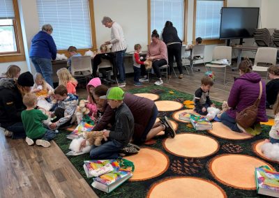 Children and adults engaged in craft activities on a colorful carpet in the YS Activity Room at the library.