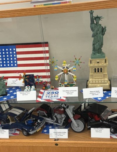 Display case featuring LEGO models including the American flag, Statue of Liberty, and vehicles, arranged with labels indicating each model's name and piece count.