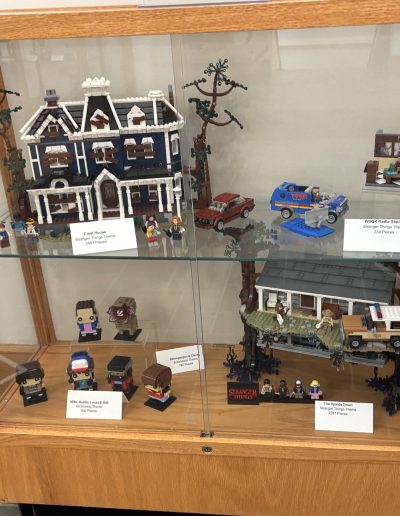 A display case showcasing various LEGO sets from "Stranger Things," including the Creel House, WSKG Radio Station, and The Upside Down, along with character figures.