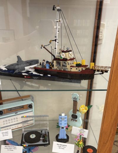 Display case showing LEGO models, including a "Jaws" ship with a shark and minifigures, a retro radio, a turntable, and a blue ukulele with flowers.
