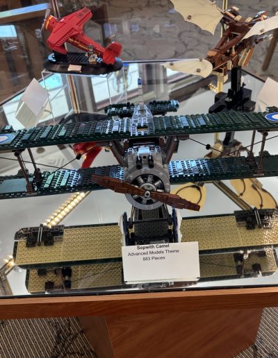 A display case showcasing three model airplanes: a green Sopwith Camel in the foreground, a red biplane to the left, and a light-colored glider to the right, all made from LEGOs.