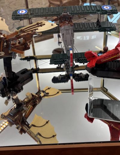 A display of LEGO model airplanes, including a flying machine with fabric wings, a red racing plane, and a green biplane, on a reflective surface.