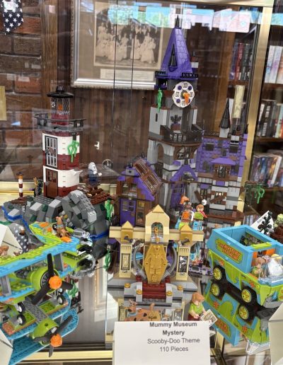 A display of LEGO "Scooby-Doo" themed sets, including a haunted castle, a lighthouse, a plane, and the Mystery Machine, all labeled with information cards.