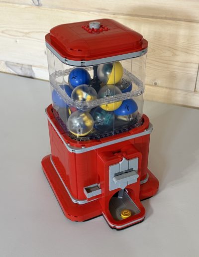 A red gumball machine made of LEGOs, filled with colorful spheres, with a coin slot at the bottom.