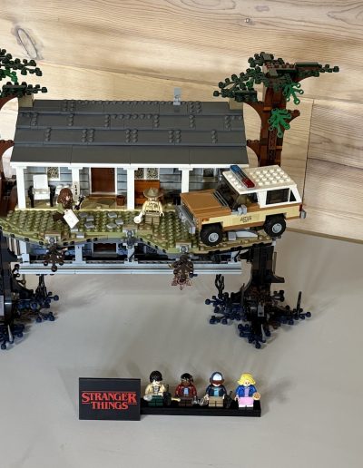 LEGO model of a "Stranger Things" house elevated on dark structures, with a police vehicle on grassy terrain and four character figures in front.