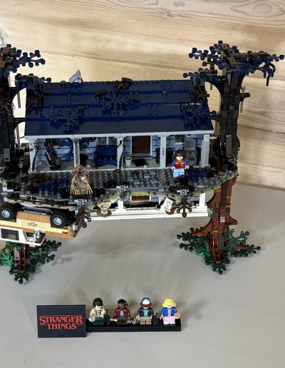 A Lego treehouse from "Stranger Things" with character figurines and a sign, displayed on a light wooden surface.