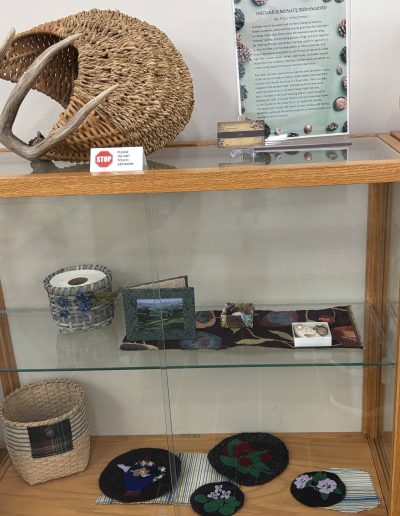 Museum display case featuring woven baskets, an antler-adorned basket, textile art, a decorated ceramic vessel, and an artist information poster.