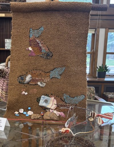 Brown woven textile artwork with colorful fish appliqués and assorted shells, buttons, stones, and found objects displayed in a glass case.
