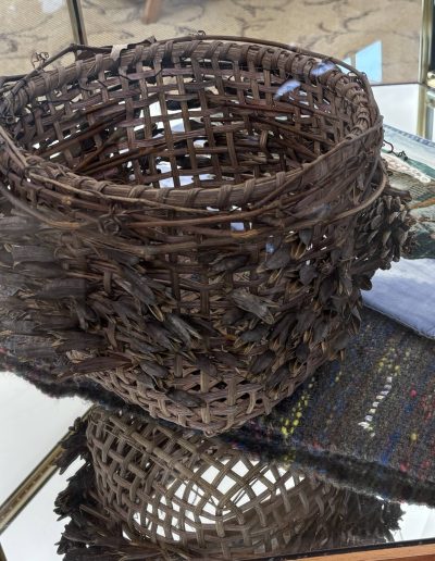 Large oval woven basket covered with layered dark leaf-like material, displayed in a glass case.