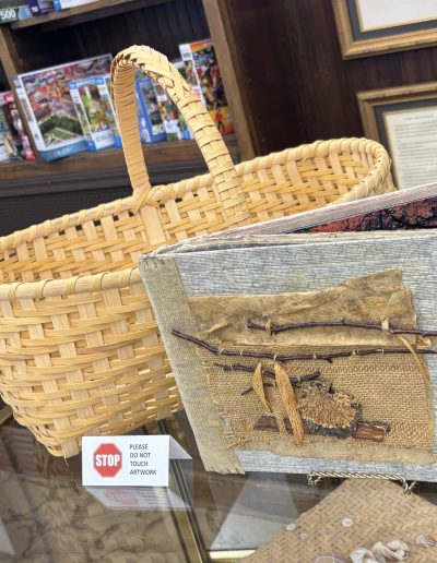 Museum display featuring a large woven basket, mixed-media artwork, and a “Please do not touch artwork” sign.